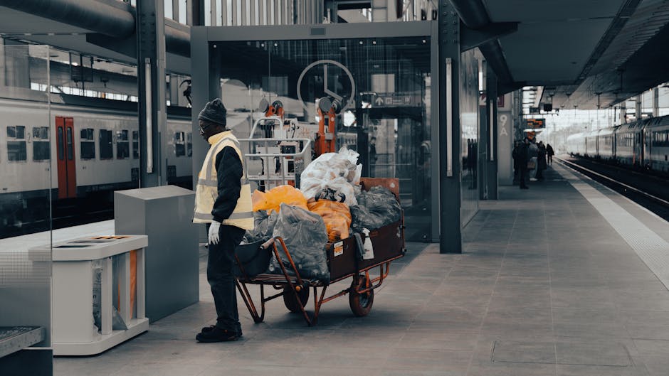 A person dressed in dark clothing and a grey beanie hat, wearing a high-visibility vest and white gloves, stands beside a red metal trolley loaded with numerous black, grey, and orange garbage bags and assorted waste items, positioned on a paved platform at a modern train station. Behind the individual, there is a glass-enclosed structure with mechanical lifts and equipment, suggesting site preparation for waste collection or handling. To the left, a white recycling bin with a partially open lid is visible, indicating waste separation efforts. In the background, multiple train carriages are parked on parallel tracks, with a few passengers walking along the platform's edge. Overhead, a metallic canopy extends along the platform, providing shelter in an industrial, urban environment. The scene appears to be captured in daytime under diffuse natural lighting, emphasizing the utilitarian aspect of waste management activity within a transportation hub. This visual context relates to private waste handling or rubbish removal services associated with public transit infrastructure or alternative rubbish collection methods, highlighting the practicalities of on-site waste clearance at a public transport facility, supported by the presence of equipment and waste containment containers typical of such settings.