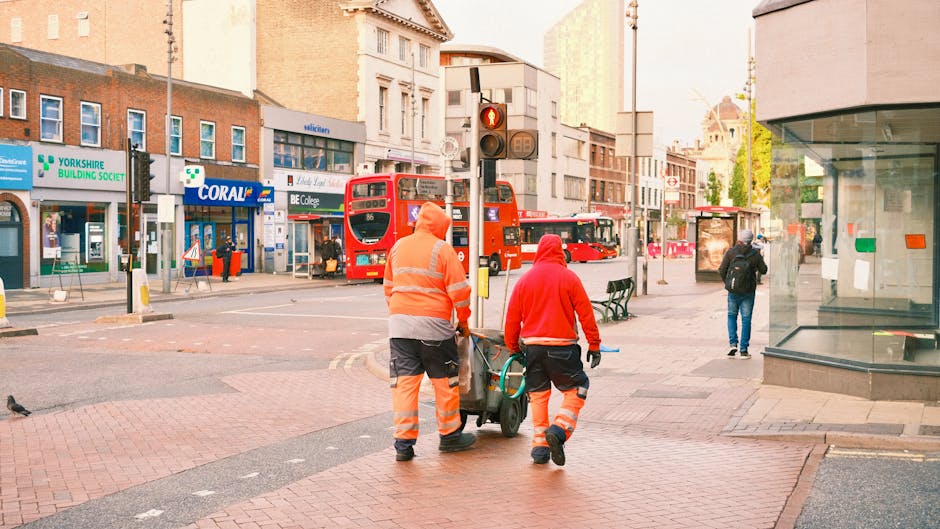 Two waste management workers wearing high-visibility orange and red safety jackets and trousers are pushing a green cart filled with waste bins or bags along a busy urban street. The scene is set on a red-brick paved sidewalk adjacent to a road with double yellow lines, featuring a traffic light showing a red signal above them. In the background, there are several double-decker buses, typical of public transport in the UK, parked or passing by, along with various shopfronts including a pharmacy, a building society, and a retail store. Pedestrians, including one person walking away with a backpack, are visible on the pavement, and a glass-fronted retail unit with a corner display window is on the right. The surroundings suggest an environment where private waste collection or rubbish removal services, such as those provided by Rubbish Removals Shepherds Bush, operate to support local waste management needs outside of standard municipal collection. The lighting appears natural, indicating daytime, with some greenery visible in the distance, adding to the cityscape context.