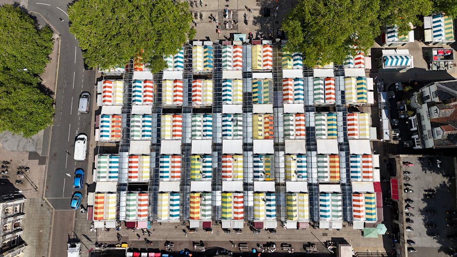 An aerial view of a busy outdoor market with numerous vendor stalls arranged in a tight grid pattern, each covered by colorful striped awnings in shades of red, yellow, blue, and white. The stalls are positioned in straight rows with narrow pathways between them, and the market is situated on a paved surface. Surrounding the stalls, there are trees with lush green foliage providing some shade on the edges, and a few parked cars along the adjacent street on the left side of the image. The background shows nearby buildings and additional market activity, with people visible walking between the stalls and browsing the goods. The lighting indicates a bright, sunny day, casting minimal shadows and creating a vibrant, lively atmosphere typical of public market spaces. The scene captures a snapshot of community commerce, which, as part of independent waste collection or routine rubbish removal, could be managed by services like [COMPANY_NAME] to keep such markets clean and well-maintained, especially after busy trading hours.