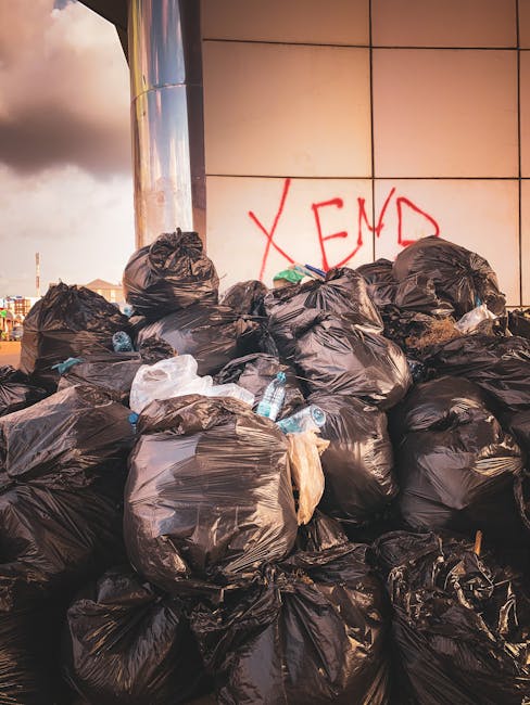 A large pile of tightly tied black plastic rubbish bags stacked against the side of a modern building with beige tiled exterior. Several discarded plastic water bottles and miscellaneous waste are partially visible protruding from some of the bags. In the background, a section of the city skyline with cloudy sky is observable, with the building surface showing graffiti in red spray paint that spells out 'XEND'. The scene appears to be outdoors on a paved surface, highlighting a situation of unmanaged waste that could relate to independent rubbish collection or illegal dumping, relevant to private rubbish removal services in the Shepherds Bush area. The lighting suggests late afternoon or early evening, casting a warm glow on the scene, emphasizing the volume of waste awaiting collection, typical of an urban setting needing professional rubbish disposal.