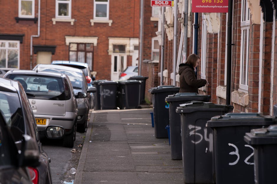 A row of black wheelie bins with closed lids, positioned along the pavement outside red-brick residential buildings, with some bins marked with white spray-painted numbers. A woman in a dark brown coat stands beside one of the bins, appearing to be either opening or closing it. The scene shows a narrow street with parked cars on the left side, including a small silver hatchback in the foreground, extending towards the background where additional houses and bins are visible. The buildings feature cream-colored window frames and stone sills, with some windows partially visible; one building has a red 'SOLD' sign in the upper right corner. The environment appears to be an urban residential area during daylight hours, with a slightly overcast sky casting diffused natural light on the scene, emphasizing the textures of the brickwork, plastic bins, and parked cars. The scene relates to waste management and private rubbish collection in a typical UK urban setting, illustrating the use of on-site waste disposal outside properties managed by services like rubbishremovalshepherdsbush.co.uk.